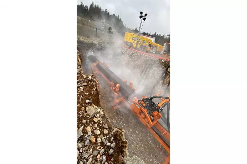 Die Druckluft aus den Atlas-Copco-Kompressoren (im Hintergrund) treibt den Schlagkolben des Imlochhammers im Inneren des Hammerbohrgeräts (groß im Bild) an, wodurch der Bohrkopf der Maschine auf die Bohrlochsohle schlägt. Durch die Schläge tritt am Kopf Luft aus, die zugleich die Sohle reinigt und das gelöste Bohrgut über den Ringraum der Maschine nach hinten und zurück an die Erdoberfläche spült. (Bild: Petzold)