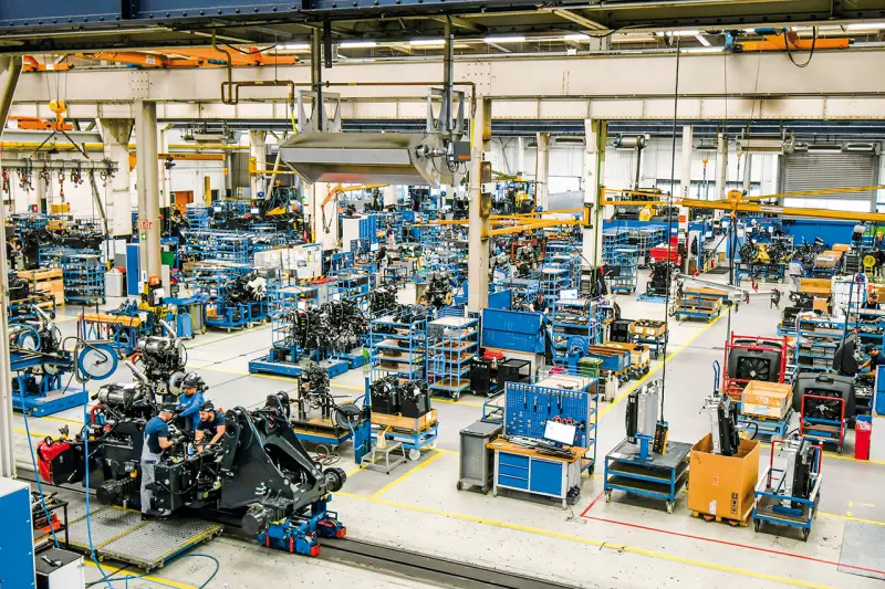 Blick auf den Fertigungsprozess der Radlader. Aus der Tiefe der Halle rücken die vormontierten Komponenten heran, die am Band zusammengebaut werden. Der Ablauf ist trotz Serienfertigung höchst flexibel. Modelle und Ausstattungen wechseln bunt durcheinander. Quelle: Treffpunkt.Bau